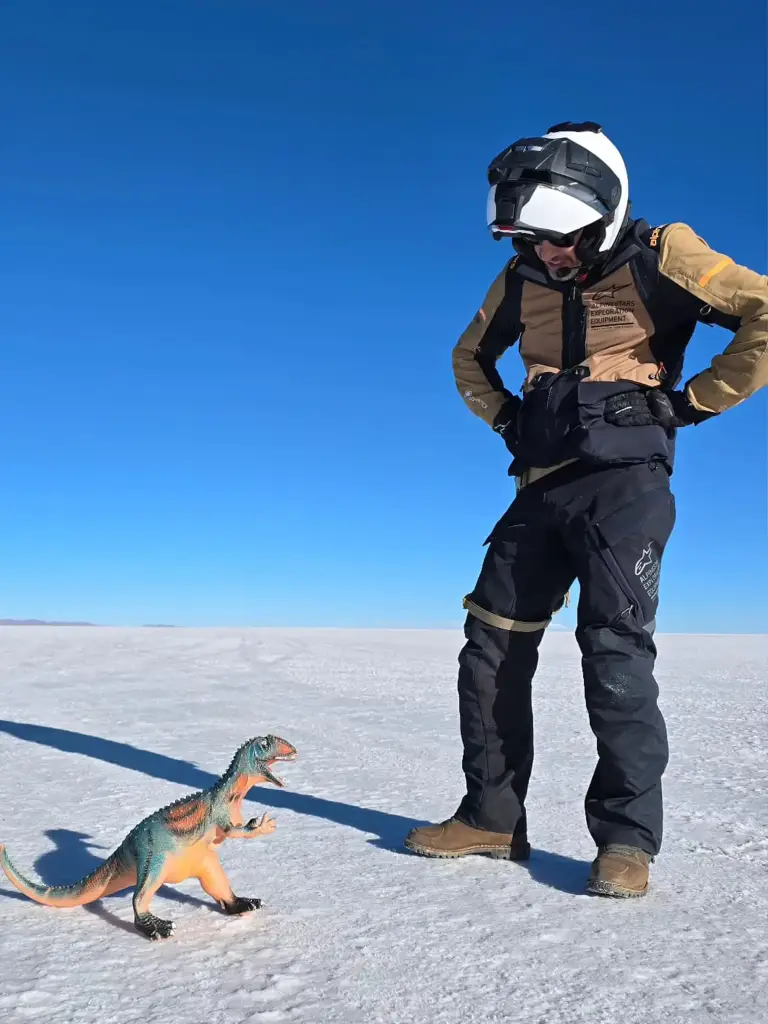 Motorcycle trip rider posing on Bolivia’s Uyuni Salt Flats next to a small toy dinosaur on the reflective white surface.