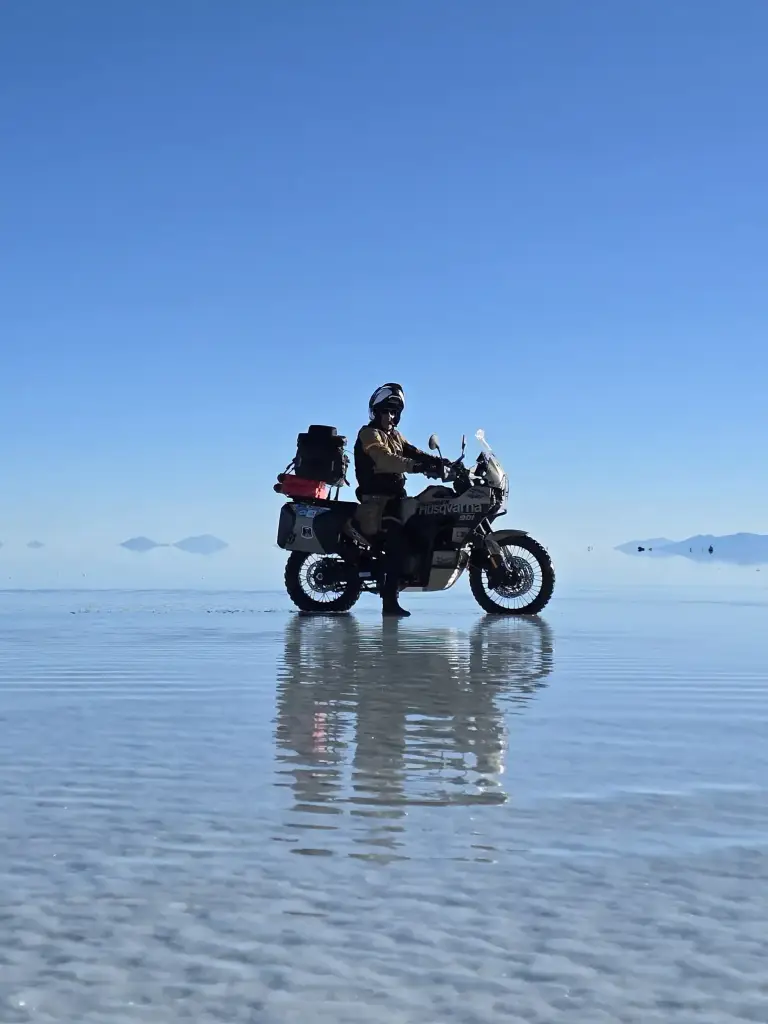Motorcyclist continuing a motorcycle trip across the mirror-like surface of Uyuni Salt Flats with distant mountains.